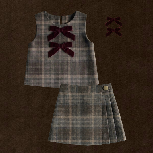 A two-piece girls' outfit set featuring a sleeveless plaid top and matching pleated skirt, both in a brown and blue plaid pattern with dark burgundy velvet bow accents on the top. The set is displayed flat on a dark brown textured background, with two matching bow accessories shown in the upper right corner.