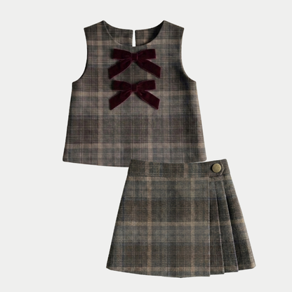 A two-piece girls' outfit set featuring a sleeveless plaid top with two dark burgundy velvet bow accents at the neckline, and a matching pleated plaid skirt with a single gold-tone button, shown on a light gray background.