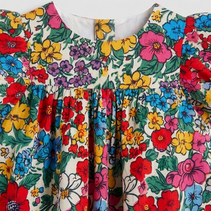 A close-up of a colorful floral print fabric, likely a children's dress or blouse, featuring a cream background with vibrant red, yellow, blue, pink, and purple flowers with green leaves.