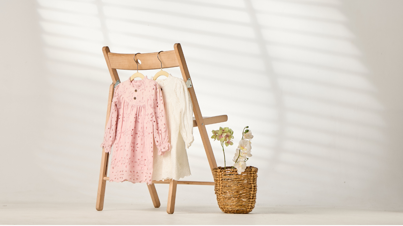 Wooden chair with a pink dress and white coat on it, next to a woven basket with a plant against a white background.